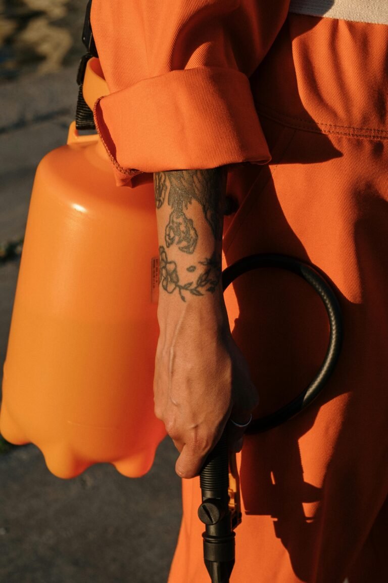 Person in orange coveralls holding a plastic sprayer, focus on tattooed arm.